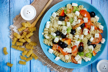 Greek salad with fresh vegetables, feta cheese, pasta and black olives on blue wooden background top view