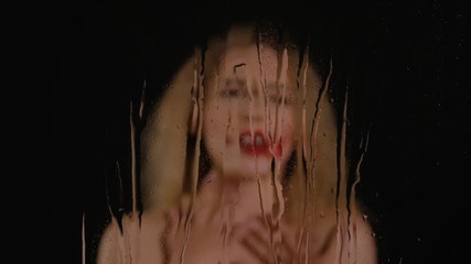 Blurred young curly blonde woman sings and touches her shoulders behind a glass with flowing drops of water. Sexy lady in a wet and rainy window on a black background