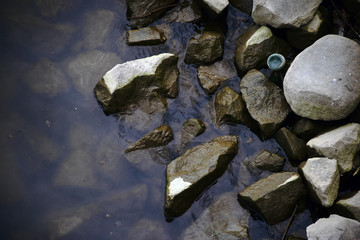 Die Uferzone eines Flusses mit Steinbrocken und Feldsteinen im Wasser sowie einer Flasche, die dazwischen klemmt.