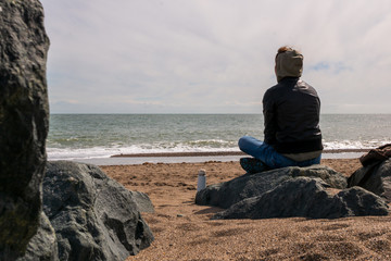 Young woman relaxing and meditating at the seashore on a rocky beach. Moody winter day seascape in Greystones, Wicklow, Ireland.