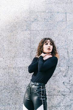 Attractive young female in stylish outfit choking herself and looking away while standing near building wall on city street