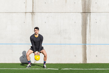 Young athletic male lifting heavy kettlebell on grass near grey wall in sunny day