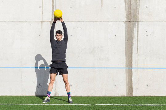 Side view of young athletic male tossing heavy training ball near tire and grey wall