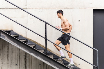 Side view of young shirtless male going up on stairs near grey wall in gym
