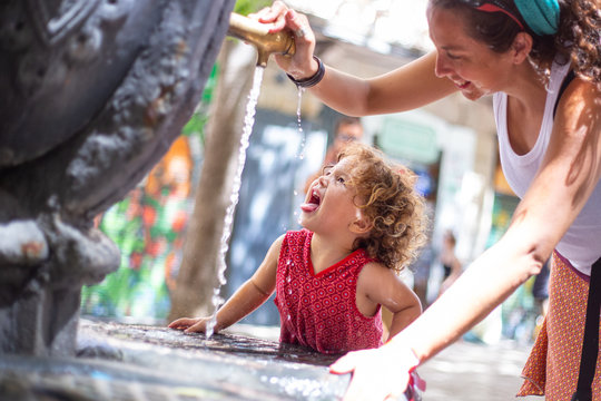 Happy mother pouring water from drinking fountain for funny girl with opened mouth on street in summer
