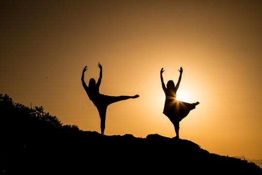 Young ballerinas with upped legs and hands dancing on hill in obscurity at sunset