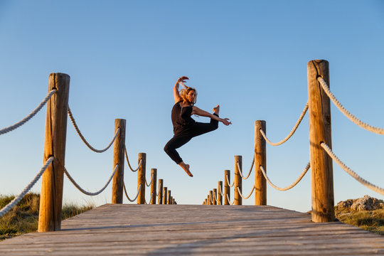 Young ballerina in black wear with upped leg in air on footbridge and blue sky in sunny day