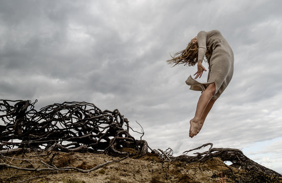 From below young ballerina in black wear with upped hands in air near dry branches and blue sky in clouds