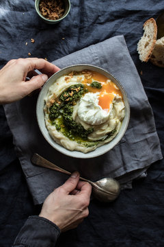From above crop hands of human holding bowl with cauliflower puree with pesto and eggs on wrinkled blue textile background