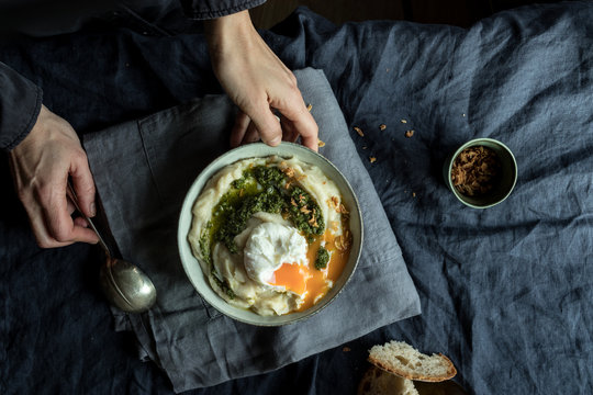 From above crop hands of human holding bowl with cauliflower puree with pesto and eggs on wrinkled blue textile background
