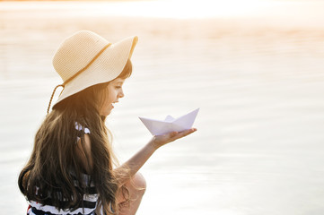 Cute little girl in hat with paper boat on the beach.