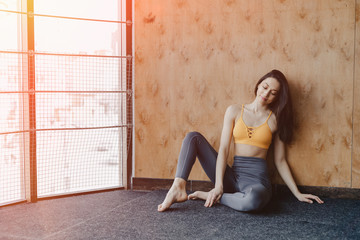 Naklejka premium Young attractive fitness girl sitting on the floor near the window on the background of a wooden wall, resting on yoga classes