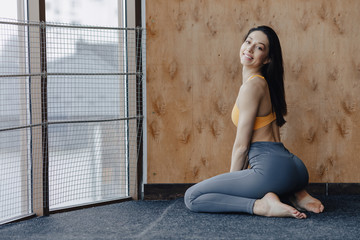 Fototapeta premium Young attractive fitness girl sitting on the floor near the window on the background of a wooden wall, resting on yoga classes