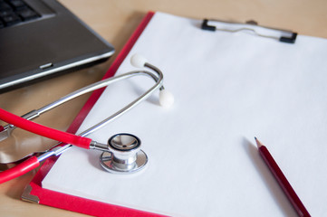 Red stethoscope and red pencil on a red clipboard on a table. Near laptop. Medical device....