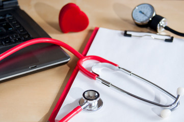 Red stethoscope, tonometer on a red clipboard. Near laptop and heart. Medical device. Treatment, health care. Heart examination. Studying the pulse.