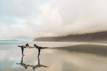 Side view of two barefoot females in sportswear performing exercise on wet sand near sea on overcast day