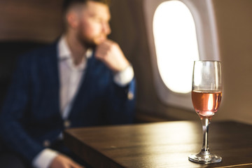 A young successful businessman in an expensive suit sits in the chair of a private jet with a glass...