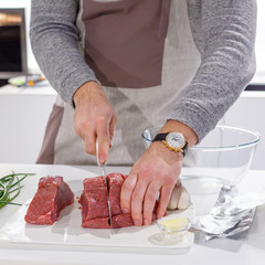 Young man cooking at home and preparing meat stake from pork or beef or lamb, in white modern light kitchen. Hands of chef cutting steak meat with knife for preparing dinner