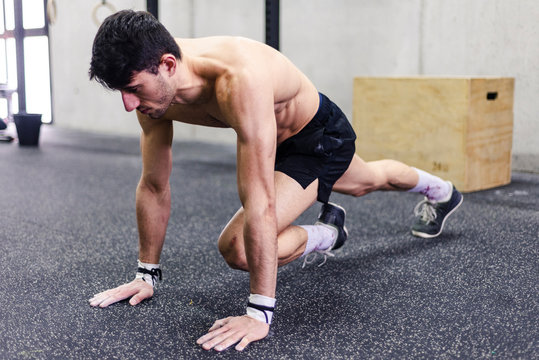 Young shirtless male doing plank and stretching legs near grey wall in gym