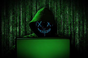 hacker bwith glowing mask behind notebook laptop in front of green source binary code background internet cyber hack attack computer concept