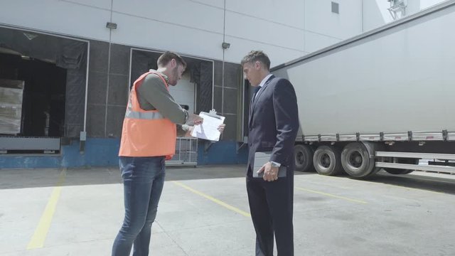 Medium shot of warehouse worker and businessman discussing over a clipboard near lorry