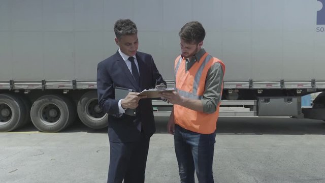 Medium shot of warehouse worker and businessman signing document and shaking hands with each other while standing near lorry
