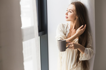 Attractive young girl in night clothes standing by the window with a mug of coffee