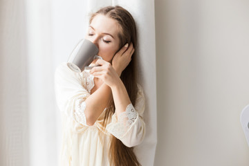 Attractive young girl in nightwear stands at the window and drinks coffee. Morning mood