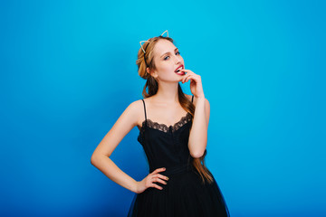 Beautiful young woman at party, masquerade, carnival, thoughtfully looking and biting finger. Wearing elegant black dress, cat ear headband with diamonds, manicure with gold polish. Blue background.