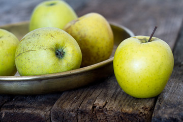 Fresh yellow-green apples lie in an old-fashioned brass plate on an old, textural, rustic table, soft focus.