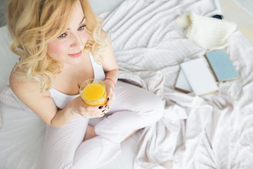 Obraz na płótnie Canvas Attractive blonde woman sits on the edge of the bed at home and holds a stocker with orange juice in her hand. Morning mood and vigor