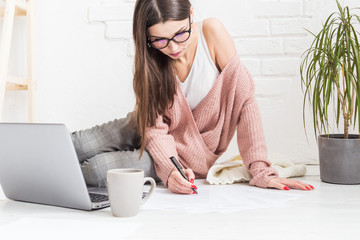 Young woman sits on the floor in a Scandinavian apartment interior with a laptop, studying law,...