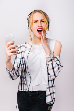 Portrait Of Young Stylish Emotional Girl In Trendy Casual Clothes Holding Hand Near Face And Reading Message. Cute Blonde With IPhone. Exciting Face. Surprised Opened Mouth. Isolated.