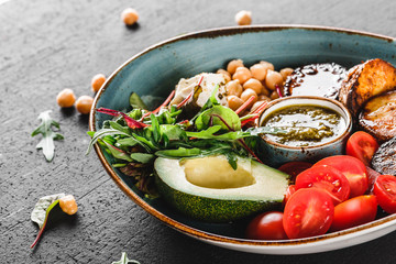 Healthy Buddha bowl dish with avocado, tomato, cheese, chickpea, fresh arugula salad, baked potatoes and sauce pesto in black background. Dieting food, clean eating.