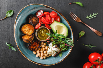 Fototapeta na wymiar Healthy Buddha bowl dish with avocado, tomato, cheese, chickpea, fresh arugula salad, baked potatoes and sauce pesto in black background. Dieting food, clean eating, top view, flat lay, copy space