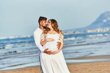 Young man hugging his pregnant wife in white dress on a beach