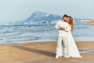 Fototapeta na wymiar Young blonde bride in white wedding dress embraces groom on the beach