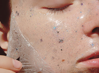 Young woman with healthy white pale skin removes purifying transparent peel off mask with shiny sparkling star glitter from her face. Trendy cosmetic beauty procedure.Skin care concept.
