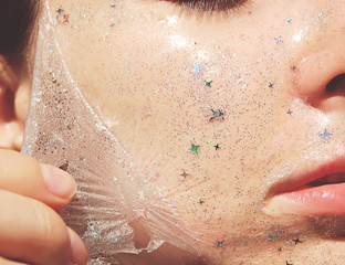 Young woman with healthy white pale skin removes purifying transparent peel off mask with shiny sparkling star glitter from her face. Trendy cosmetic beauty procedure.Skin care concept. 