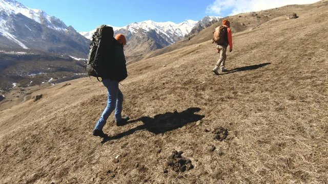 Two tourists photographers with backpacks in hats and sunglasses go up the hill on the yellow grass with cameras in their hands against the background of snowy mountains. slow motion