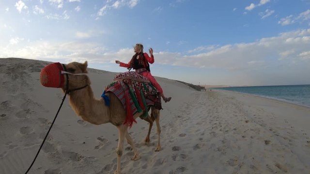 Inland sea is a major tourist destination for Qatar. Freedom woman riding camel on beach at Khor al Udaid in Persian Gulf. Caucasian blonde tourist enjoys camel ride in Middle East, Arabian Peninsula.
