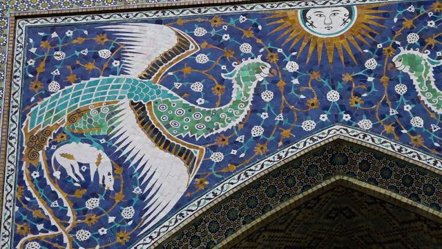 Handheld, panning, exterior, low angle, medium close up shot of a mural with a sun and a phoenix on the front wall of the Nadir Divan-Begi madrasah.