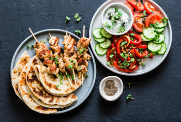 Mediterranean style lunch table - turkey skewers, flatbread, tomatoes, cucumber salad, baked sweet pepper, yogurt herb sauce  on a dark background, top view