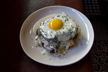 Croque madame on white plate against dark brown leather background.  It's a variation of the The croque monsieur, the classic French ham and cheese sandwich covered in cheesy bechamel.