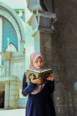 Young malay muslim women reading the holy quran while leaning on a pillar outside a mosque.
