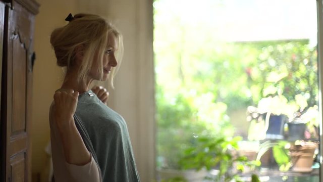 Medium shot of a mature woman standing in the bedroom holding top against her chest and smiling