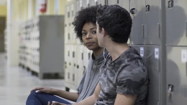 Medium shot of teenage boy and girl sharing earphones and listening to music while sitting in locker room