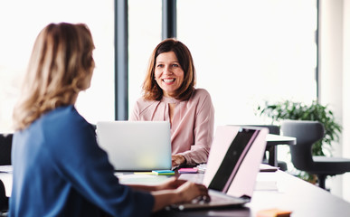 Two female business people with laptop sitting in an office, talking.