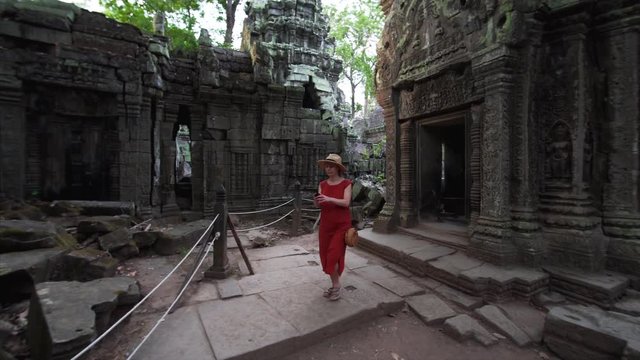 Young woman in red dress is taking photos of famous Ta Prohm temple ruins and huge banyan roots on them. Angkor Wat complex. Cambodia. UNESCO inscribed Ta Prohm on the World Heritage List in 1992