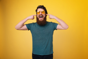 Photo of handsome guy screaming and putting fingers in ears, yellow isolated background